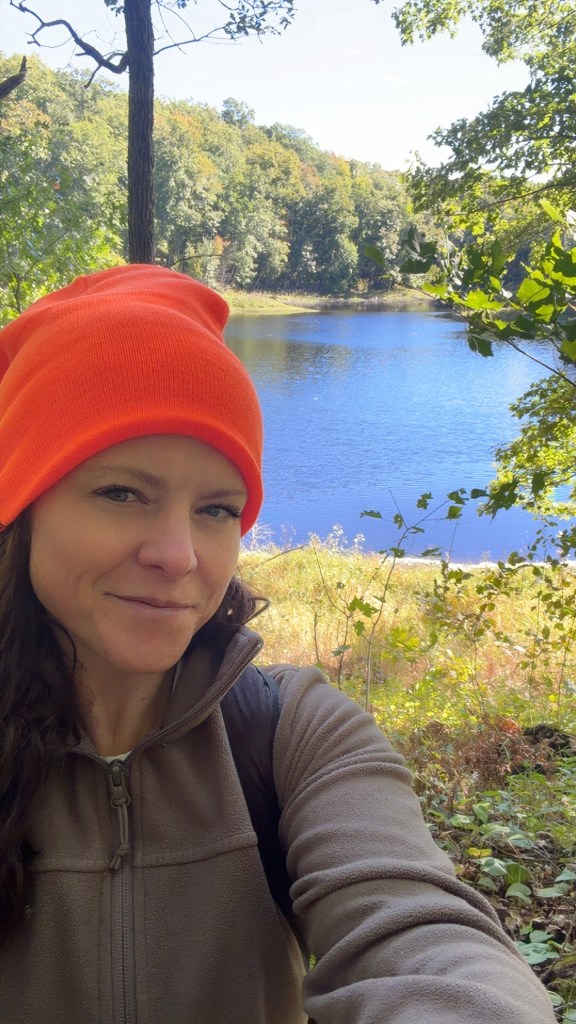 Katrina in an orange hat beside a lake and wooded shoreline.